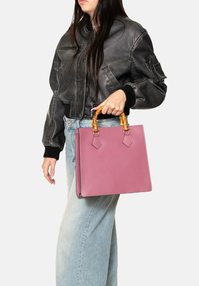 Roze lederen handtas met een gestructureerde vorm, voorzien van een bamboehendel en hoekige accenten, gedragen naast een zwarte bomberjack en lichte jeans.