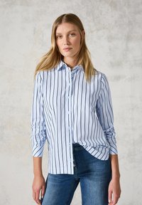 Light blue button-up shirt with navy vertical stripes, tailored fit, long sleeves, and a curved hem, worn with dark blue jeans.