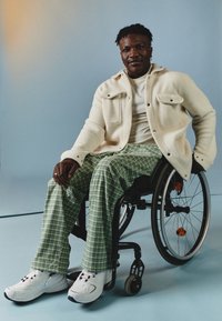 Witte fleece jack over een wit T-shirt, groene geruite broek en witte sneakers. De persoon zit in een rolstoel tegen een blauwe achtergrond.