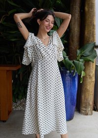 Femme souriante, les bras levés, portant une robe blanche à pois noirs, debout à l'intérieur près d’un grand pot bleu et des plantes tropicales.