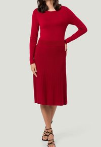 Robe rouge côtelée avec des manches longues et une taille cintrée, se transformant en une jupe plissée. Le tissu a une texture lisse et un éclat subtil.