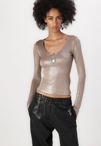 Metaalachtige top met lange mouwen in taupe, met een ronde halslijn en knoopdetail. Gecombineerd met zwarte geplooide pantalons met een elastische tailleband.