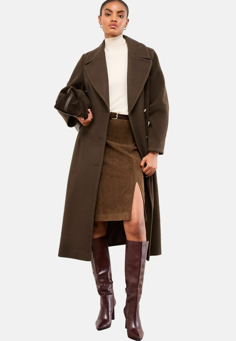 Abrigo de lana marrón con diseño de doble botonadura, suéter de cuello alto color crema, falda de ante marrón con abertura lateral y botas de charol burdeos hasta la rodilla.