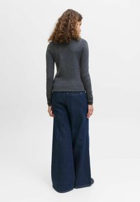 Žena s kudrnatými vlasy, oblečená v přiléhavém šedém dlouhém rukávu a širokých temně modrých džínách, stojí zády k nám.