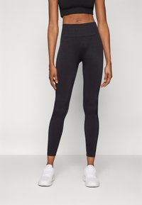 Fekete, magas derékú, rugalmas anyagból készült leggings, sima textúrával és karcsúsított szabással, fehér sportcipővel párosítva.