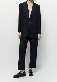 Blazer oversize bleu marine avec un col en V profond, associé à un pantalon assorti. Mocassins à plateforme noirs brillants avec des détails en métal doré.