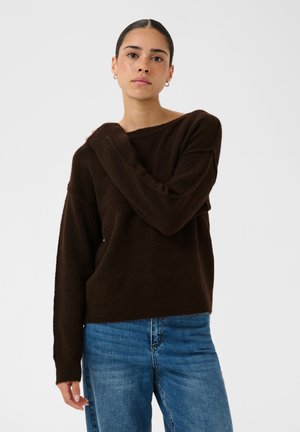Jeune femme aux cheveux foncés en chignon, portant un pull marron foncé et un jean bleu, posant avec une main sur son épaule opposée.