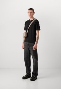 Zwart T-shirt met korte mouwen, donkere jeans en zwarte schoenen. Kleine beige schoudertas met een ritssluiting. Model met een zichtbare tatoeage op de arm.