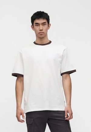 Ung mand med kort sort hår iført en hvid t-shirt med mørk kant ved halsudskæring og ærmer, står mod en ensartet lys baggrund.