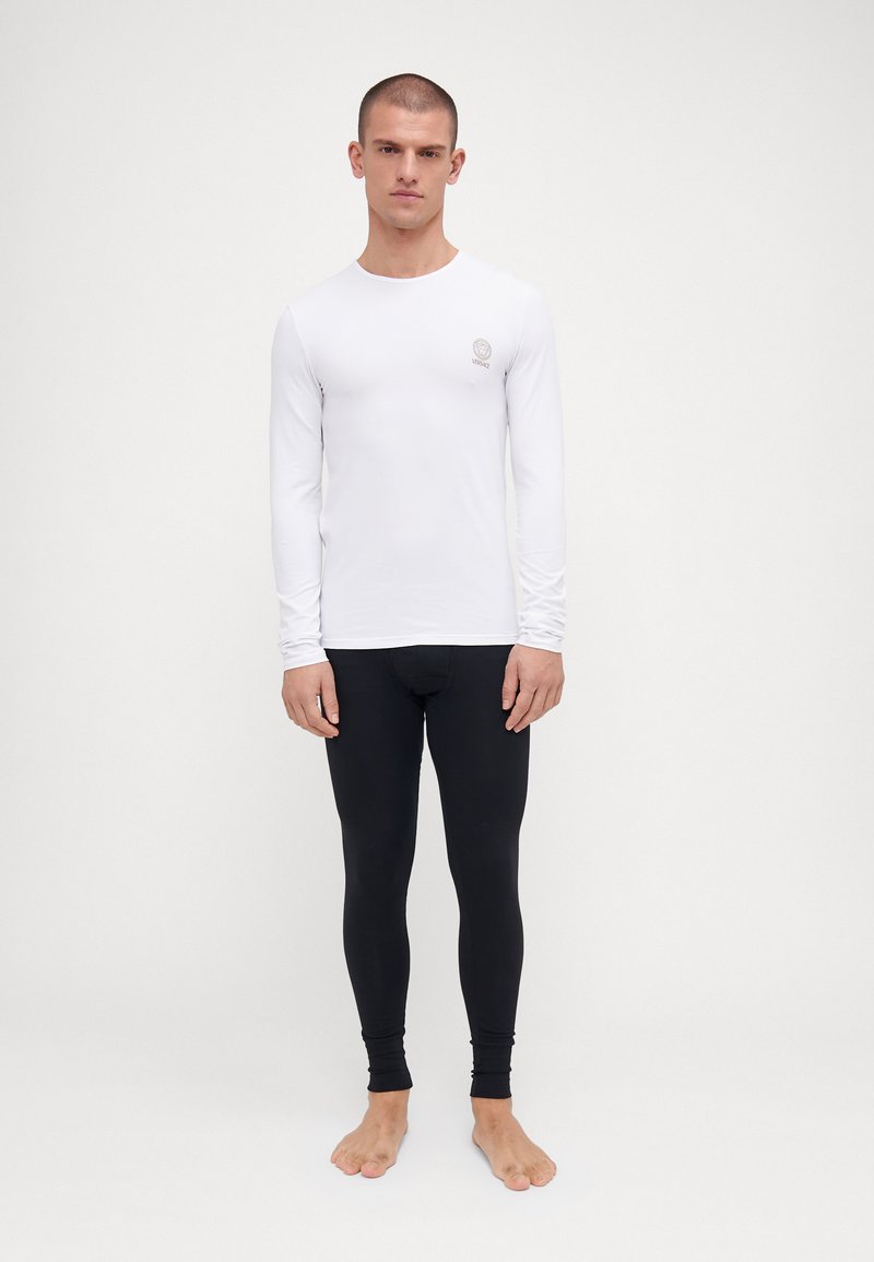 Weißes Langarmshirt mit einem Rundhalsausschnitt und kleinem Logo, kombiniert mit schwarzen, eng anliegenden Leggings. Glatte Stoffqualität, ohne Muster oder Akzente.