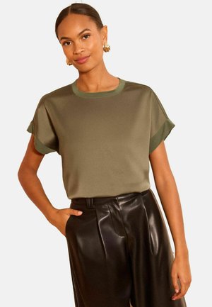 Naine, kes kannab oliivirohelist lühikeste varrukatega toppi mansettidega ja musti kõrge vöökohaga nahkpükse, poseerib ühe käega taskus.