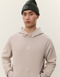 Beige hette med snøring og frontlomme, med en hvit "II" logo. Modellen har på seg en olivengrønn ribbestrikket lue. Glatt tekstur.