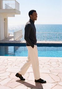 Homme portant un pull noir et un pantalon blanc marchant à côté d'une piscine extérieure avec vue sur l'océan sous un soleil éclatant.