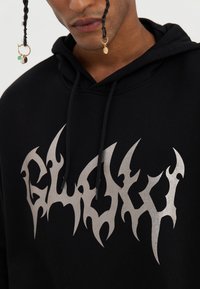Zwarte hoodie met een grote zilveren vlamachtige grafiek op de voorkant. Inclusief zwarte trekkoorden en een zachte, katoenen textuur.