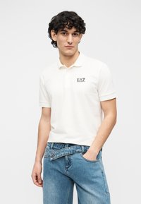 Polo color crema de manga corta, con un logo EA7 negro en el pecho izquierdo, combinado con jeans de mezclilla azul claro.