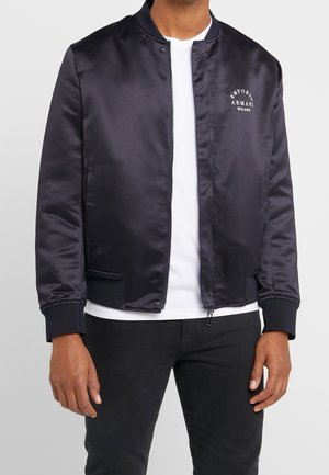 Mand iført en marineblå satin bomberjakke med "Emporio Armani Milano" logo på brystet, over en hvid t-shirt og sorte bukser.