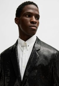 Schwarzer Pailletten-Blazer mit scharfem Rever, getragen über einem weißen Hemd mit Kragen. Die strukturierte Oberfläche reflektiert das Licht und erzeugt einen schimmernden Effekt.