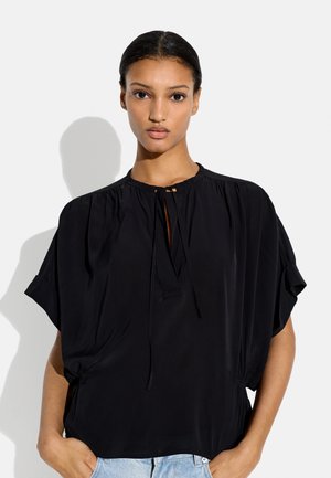 Kobieta z włosami zaczesanymi do tyłu nosi luźną czarną bluzkę z wiązanym dekoltem i krótkimi rękawami, dobraną do jasnoniebieskich dżinsów.