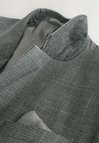 Jachetă gri cu model și guler înalt. Prezintă o textură moale și un batiste de buzunar gri deschis vizibil în buzunarul de la piept.