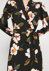 Robe portefeuille noire avec grand imprimé floral blanc et jaune, manches longues bouffantes et ceinture nouée à la taille.
