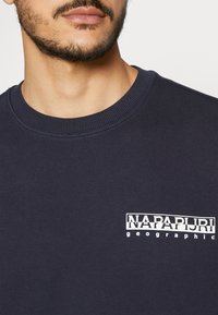 Felpa blu navy con collo a girocotolo a costine e logo bianco "NAPAPURI geographic" sulla parte sinistra del petto. Tessuto dalla consistenza liscia.