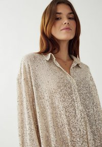 Blizganti smėlio spalvos palaidinė su blizgučiais, apykakle ir sagų uždarymu. Laisvas siluetas ir lygus paviršius, su švytinčia danga ir vertikalia raštų struktūra.