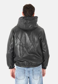 Uomo che indossa un giubbotto in pelle nera con cappuccio e jeans in denim azzurro chiaro, visto da dietro su uno sfondo bianco semplice.