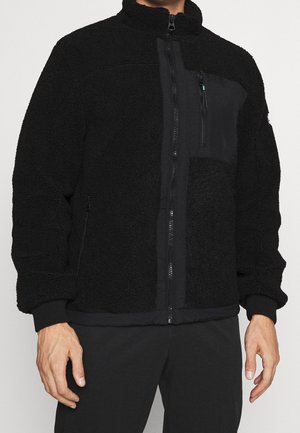 Homme portant une veste polaire noire zippée avec un col montant et une poche poitrine zippée, associée à un pantalon noir.