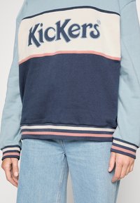 Sweat-shirt avec blocs de couleurs bleu, blanc et rose, poignets et ourlet rayés, et logo "Kickers" sur la poitrine, porté avec un jean bleu clair.