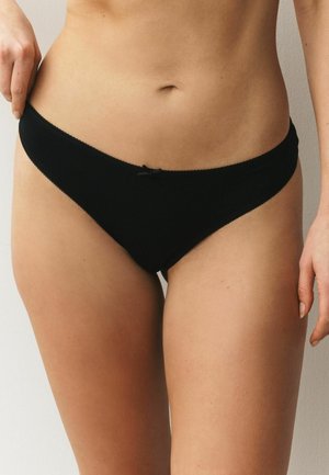 Frau trägt schwarze Baumwollbikini-Unterwäsche, steht mit einer Hand auf der Hüfte vor einem schlichten hellen Hintergrund.