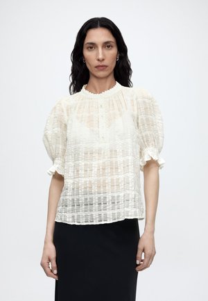 Kobieta o długich ciemnych włosach nosi białą przezroczystą, teksturowaną bluzkę z bufkami na rękawach oraz czarną spódnicę na jednolitym jasnym tle.