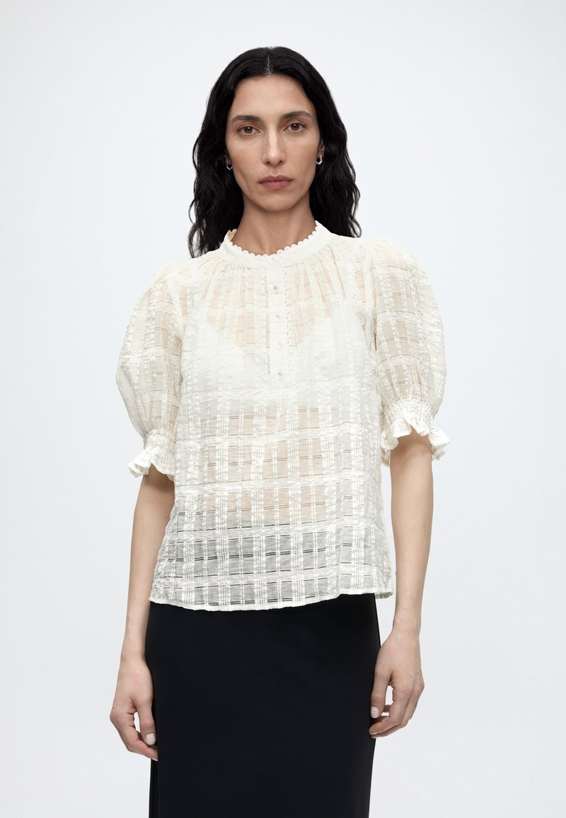 Γυναίκα με μακριά σκούρα μαλλιά φοράει λευκό διαφανές υφασμάτινο μπλουζάκι με φουσκωτά μανίκια και μαύρη φούστα πάνω σε απλό ανοιχτό φόντο.