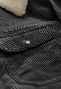 Tejido de pana negra con rayas verticales, que cuenta con un bolsillo parche con un botón de presión plateado y un suave cuello de borreguillo beige.