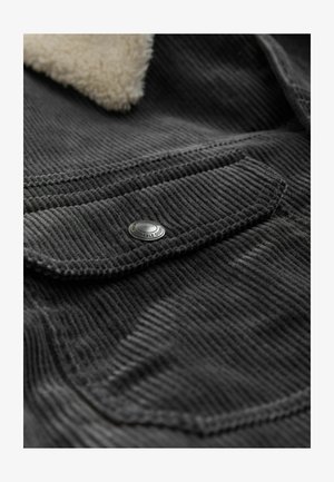 Schwarzer Cordstoff mit vertikalen Rippungen, ausgestattet mit einer aufgesetzten Tasche mit einem silbernen Druckknopf und einem weichen, beigen Schafslederkragen.