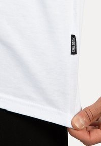 Camiseta blanca de algodón con bajo recto y abertura lateral. Presenta una etiqueta negra con "SMILOOX" cosida cerca del bajo. Textura suave.