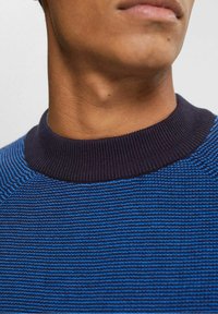 Blå och marinrandig stickad tröja med ribbad krage. Texturen är jämn med ett subtilt horisontellt mönster. Nära bild av halsringningen.