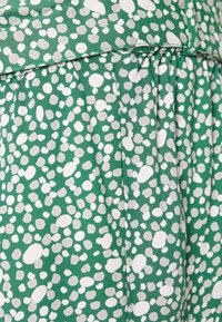 Tissu vert avec un motif de petites pierres blanches et gris clair, présentant des plis et une texture lisse. Finition avec une couture supérieure.