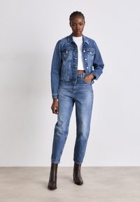 Traper jakna s ukrasima od rhinestona, u kombinaciji s trapericama visokog struka. Model nosi bijeli crop top i smeđe čizme s visokom petom.