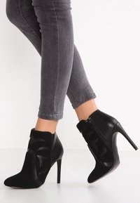 Botines negros hasta el tobillo con tacones de aguja, que combinan texturas lisas y de ante, con diseño recortado y cierre lateral con cremallera.