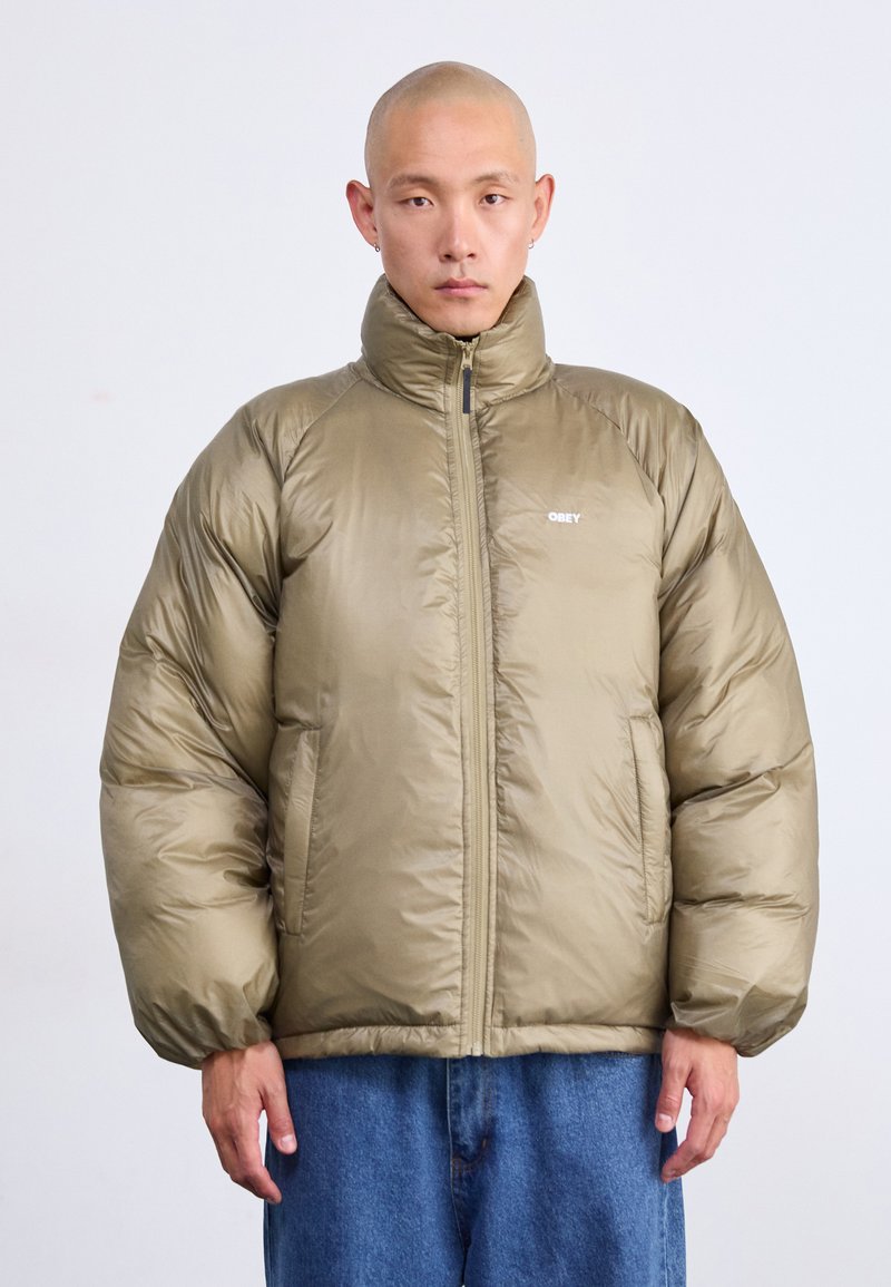 Homem com a cabeça rapada, vestindo uma jaqueta acolchoada verde-oliva com o logótipo "OBEY" e jeans azuis, de pé contra um fundo claro e simples.