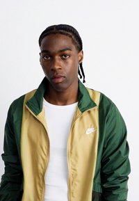 Zeleno-rjava Nike zip-up jakna z gladko teksturo, s svojim belim logotipom na levem prsnem predelu, nosi se čez belo majico.