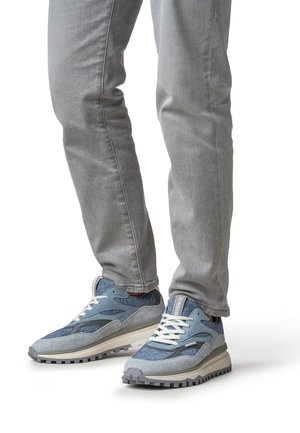 Baskets bleues avec textures de tissu mélangées, présentant une combinaison de daim et de maille, semelle grise et lacets blancs. Portées avec un jean gris clair.