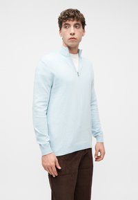 Νεαρός άνδρας που φοράει ανοιχτό μπλε πουλόβερ με φερμουάρ στο στήθος πάνω από λευκό πουκάμισο και καφέ παντελόνι από κορντόνι, κοιτάζοντας ελαφρώς στο πλάι, μπροστά σε απλό φόντο.