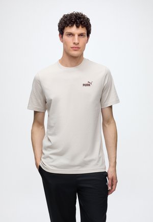 Homme aux cheveux bouclés debout, portant un t-shirt beige clair Puma et un pantalon noir avec les mains dans les poches, sur un fond blanc uni.