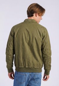 Mies selin, päällään oliivinvihreä bomber-takki ja siniset farkut, vaaleaa taustaa vasten.