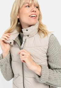 Helles beige Steppweste mit hohem Kragen, Reißverschluss und Druckknöpfen; kombiniert mit einem strukturierten grauen Strickpullover.