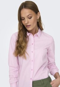Camicia button-up a righe rosa chiaro con maniche lunghe e colletto. Presenta un orlo arrotondato e bottoni discreti. Indossata con pantaloni verdi.
