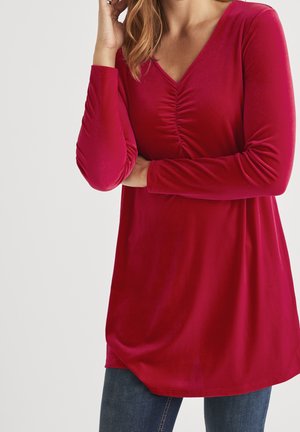 Femme portant un haut tunique rouge à manches longues avec un col en V froncé et un jean bleu, posant avec un bras croisé et une main près du visage.