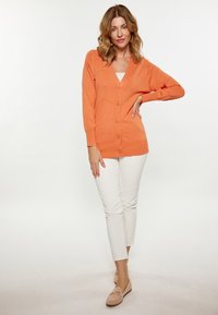 Cardigan lavorato a maglia arancione con scollo a v, chiusura a bottoni e un motivo intricato. Abbinato a pantaloni bianchi slim-fit e mocassini beige.