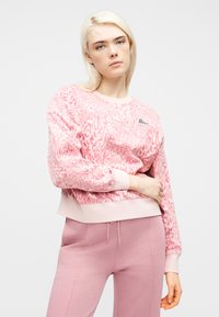 Νεαρή γυναίκα που φοράει ροζ φούτερ με animal print και ροζ παντελόνι, στέκεται με το ένα χέρι στη μέση της μπροστά σε ένα απλό λευκό φόντο.